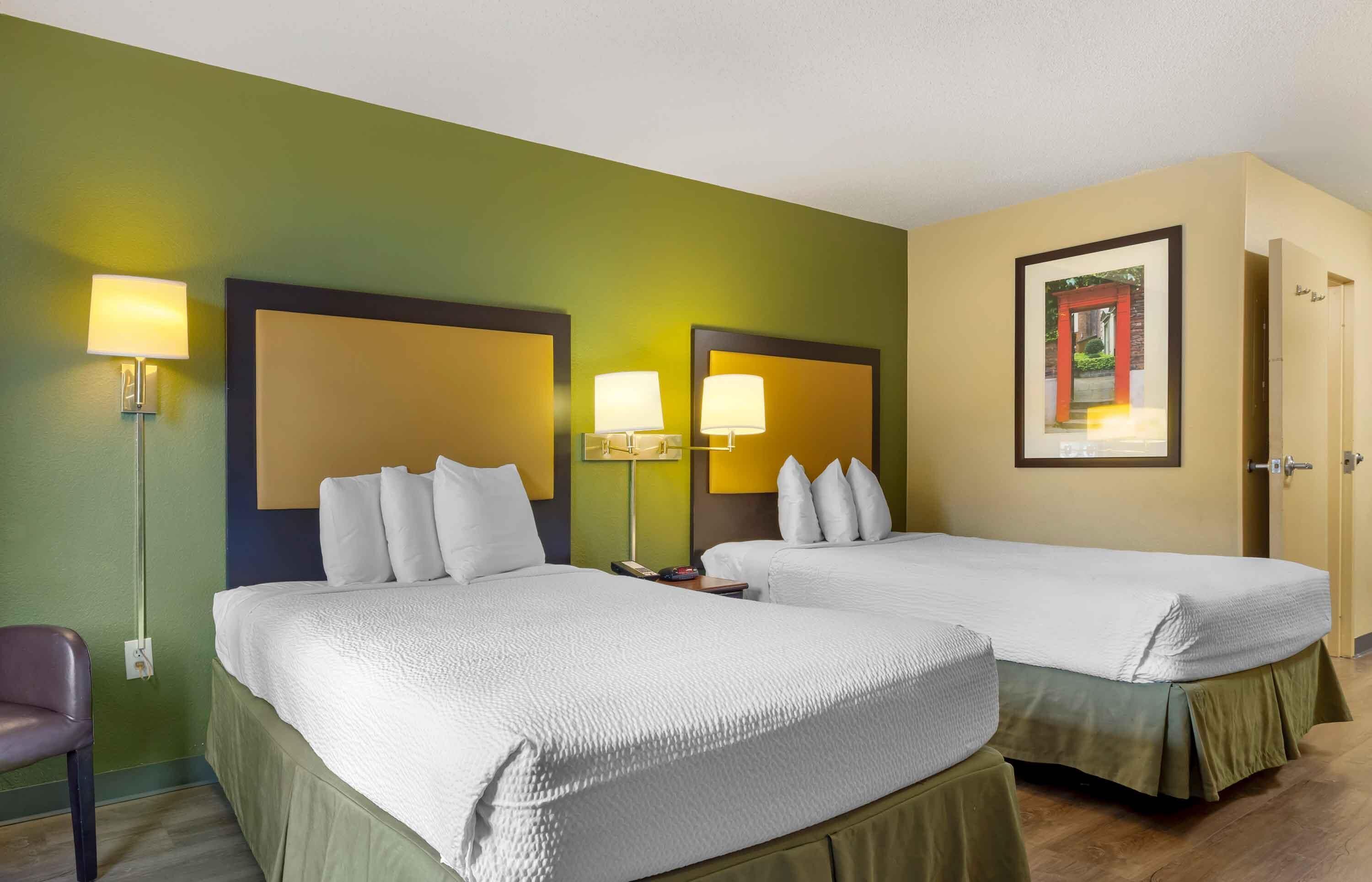 Ein Hotelzimmer mit zwei Betten, jeweils mit Kopfteil und Nachttischlampen. Ein Stuhl und eine Tür sind rechts zu sehen.