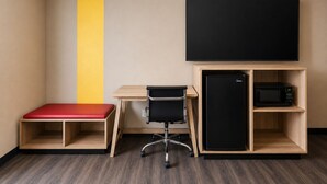 Ein kleiner Raum mit Schreibtisch, schwarzem Bürostuhl, roter Bank, schwarzem Fernseher und schwarzem Kühlschrank.