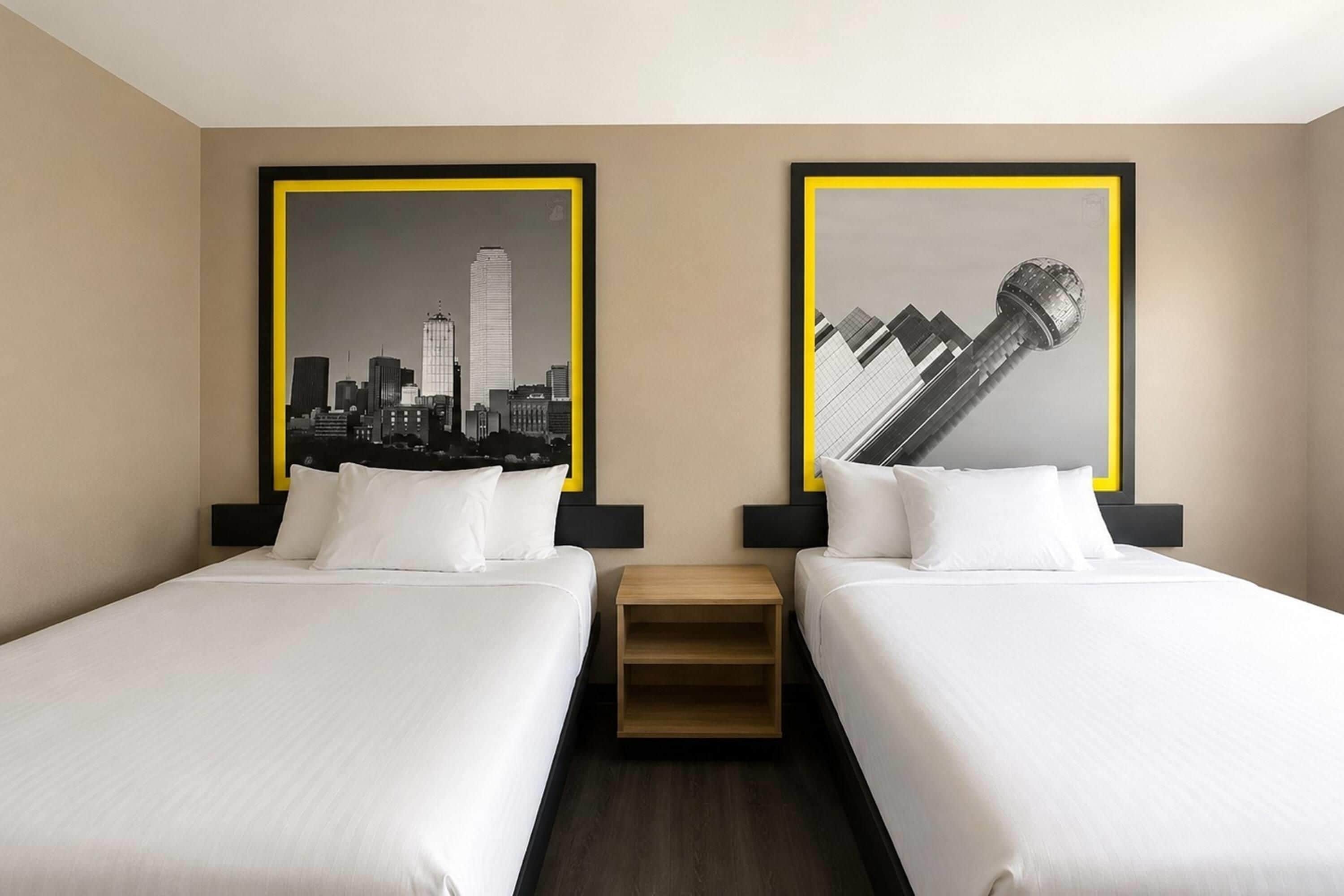 A hotel room with two beds, each with white bedding and black headboards. There are two framed black and white cityscape photographs on the wall.