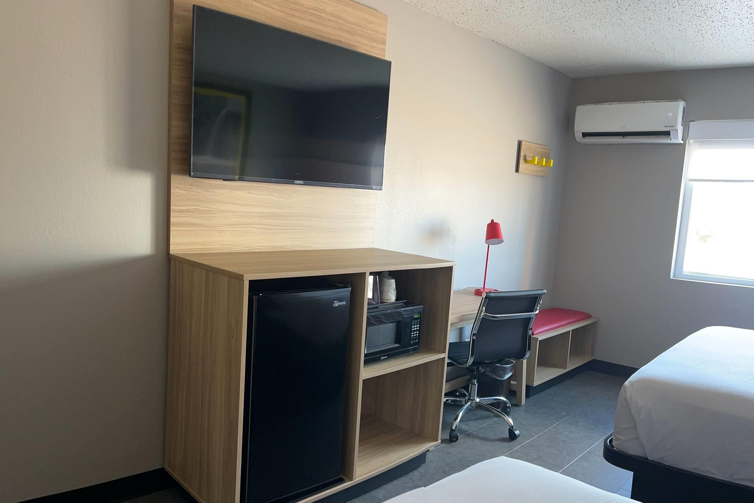 A hotel room with a flat-screen TV mounted on the wall, a small refrigerator, a desk with a chair, and a bed with a white comforter.
