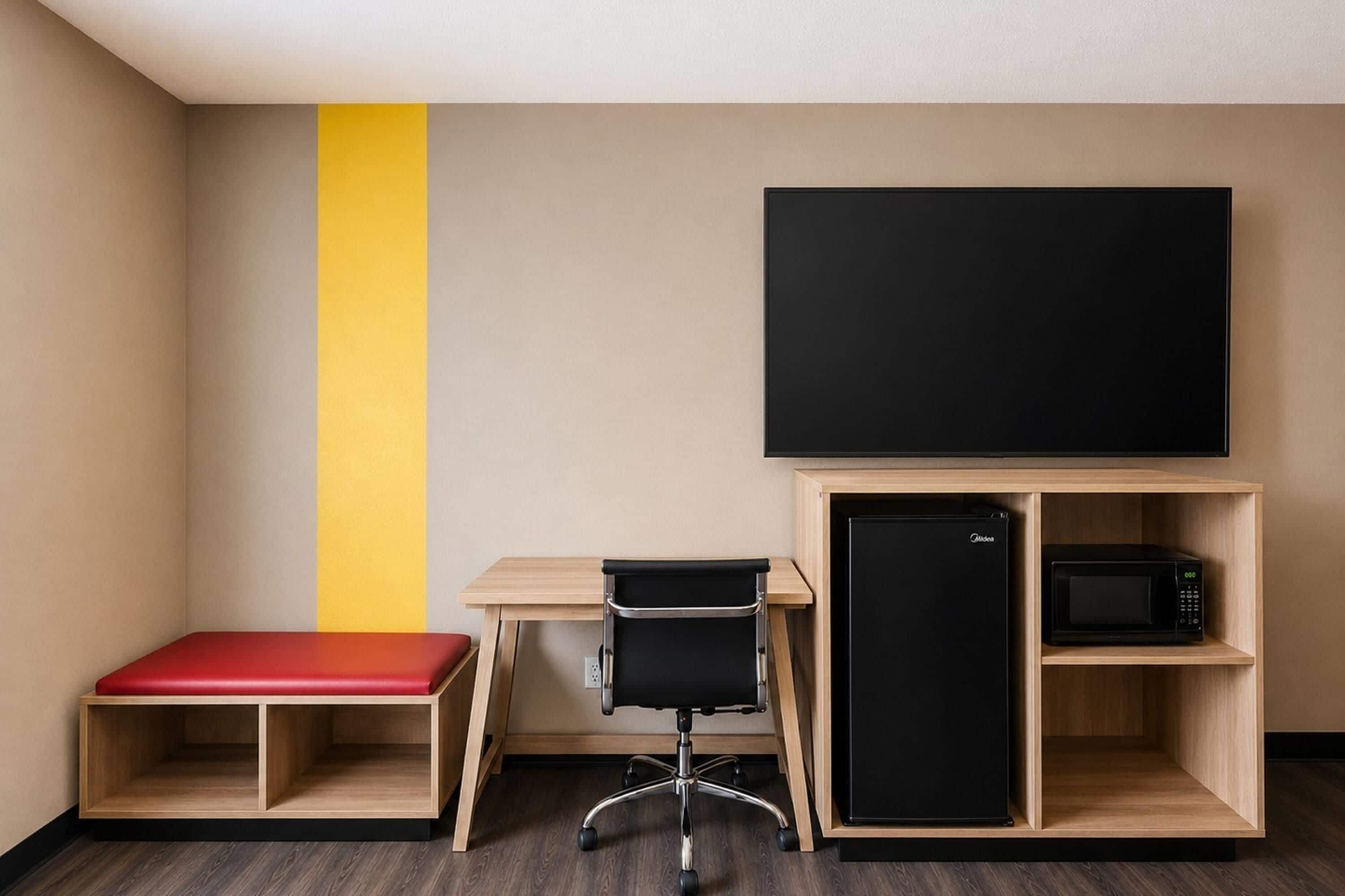 Habitación de hotel con escritorio, un banco rojo, una silla de oficina negra, un refrigerador negro y un microondas negro.