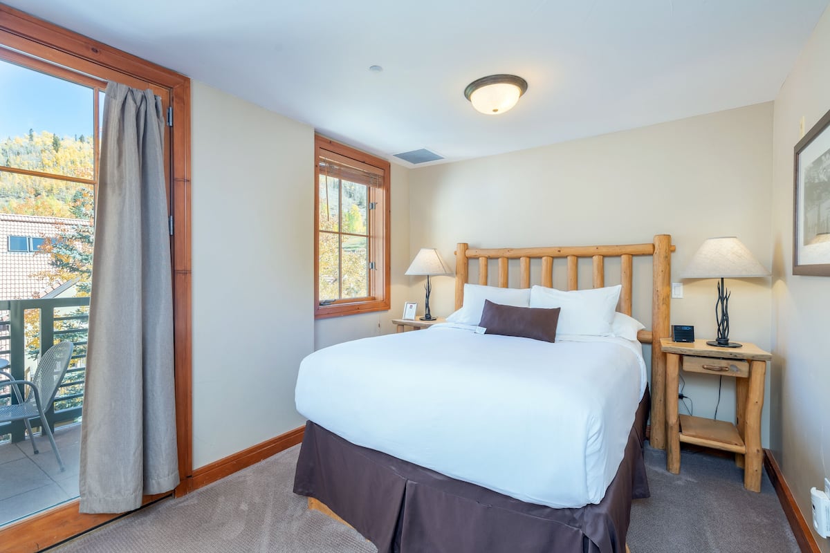 Une chambre avec un grand lit, une tête de lit en bois, deux tables de chevet avec des lampes, une fenêtre avec des rideaux et une vue sur un balcon.
