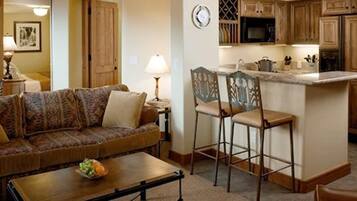 A living room with a sofa, a coffee table with fruit, and a kitchen area with wooden cabinets and a microwave.