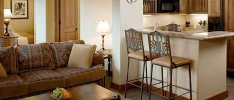 A living room with a sofa, a coffee table with fruit, and a kitchen area with wooden cabinets and a microwave.