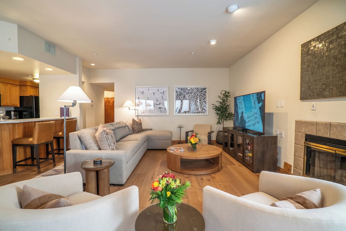 Un salon moderne comprenant un canapé, des fauteuils, une table basse et une cheminée. Il y a également un coin cuisine avec des meubles en bois et un réfrigérateur.