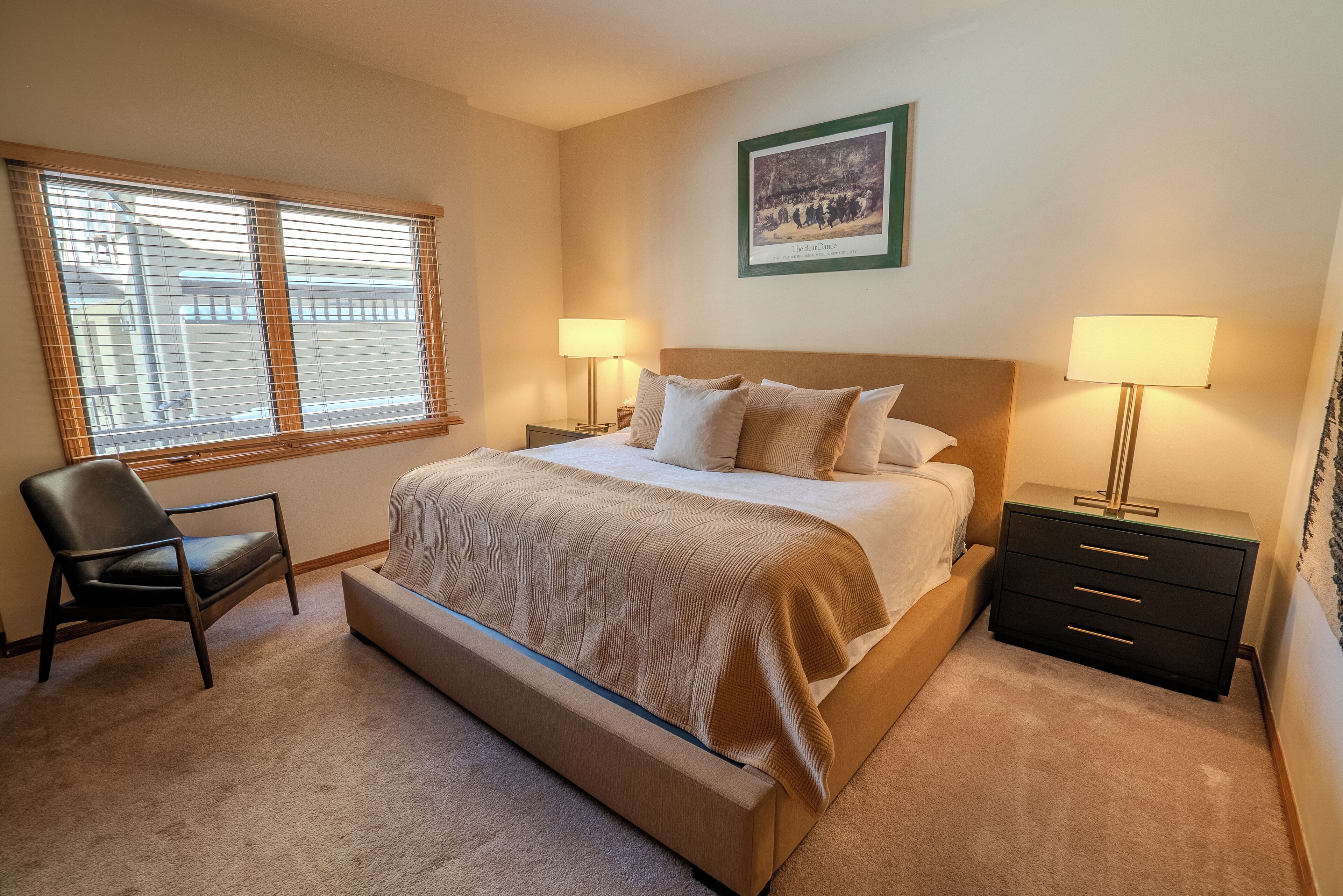 A bedroom with a bed, two bedside tables with lamps, a chair, a window with blinds, and a framed picture on the wall.