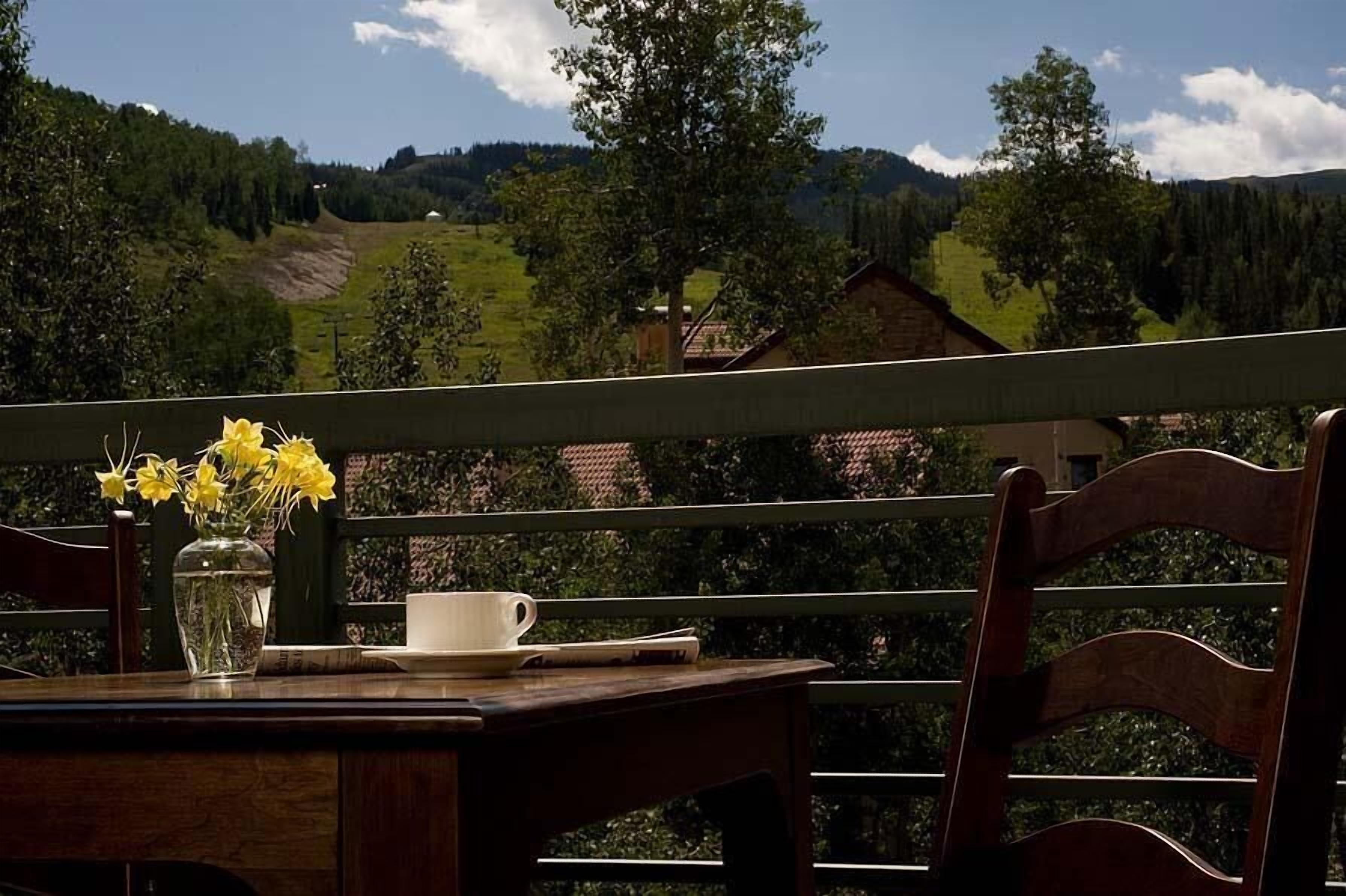 A wooden table with a vase of yellow flowers, a white cup, and a newspaper on a balcony overlooking a hilly landscape with trees and a building in the distance.