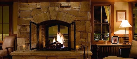 Una chimenea de piedra con fuego, un alero de madera y una zona de estar acogedora con una silla y un banco acolchado.