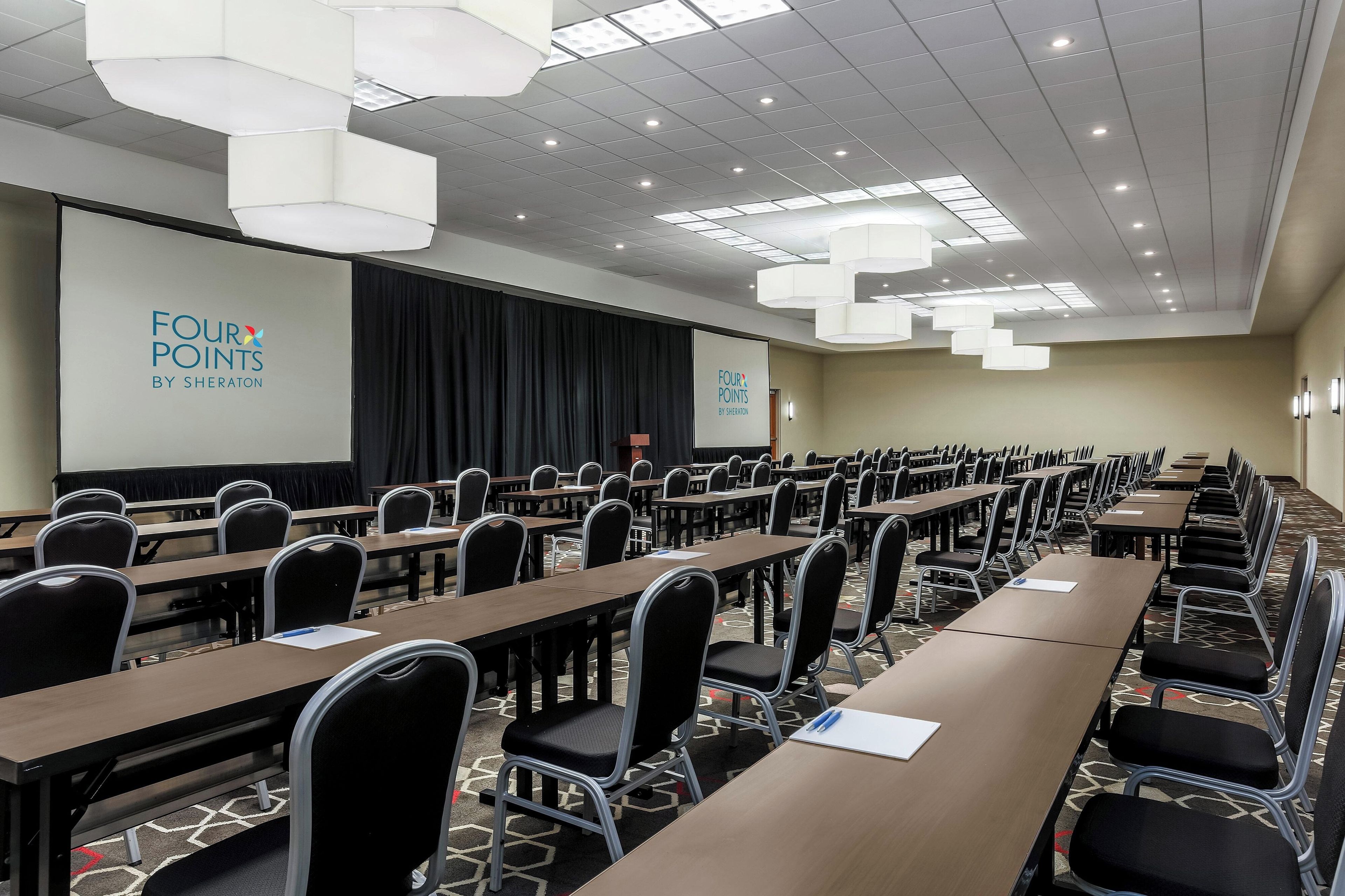 Uma sala de conferências com fileiras de mesas e cadeiras, uma tela de projetor e um palco com cortinas.
