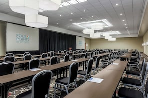 A conference room with rows of tables and chairs, a projector screen, and a stage with curtains. - Four Points by Sheraton Bentonville (Bentonville)