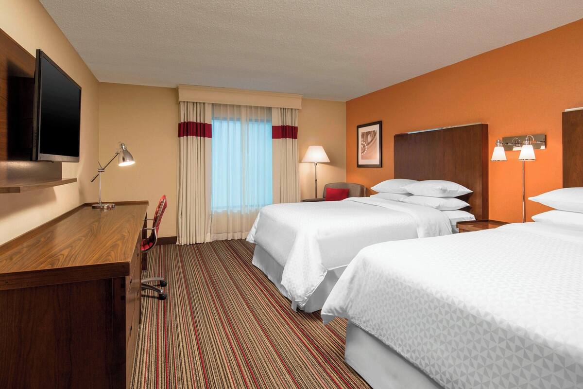 A hotel room with two beds, a desk, a TV, and a window with curtains.