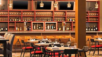 Zona de bar con varias estanterías llenas de botellas y vasos, mesas de madera con sillas tapizadas en rojo y lámparas colgantes.