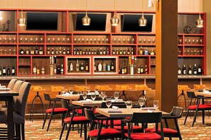A bar area with multiple shelves of bottles and glasses, wooden tables with red cushioned chairs, and hanging pendant lights. - Four Points by Sheraton Bentonville (Bentonville)