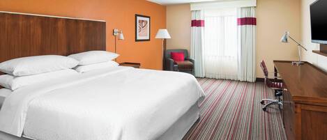 A hotel room with a large bed, a desk, a chair, a TV, and a window with curtains.