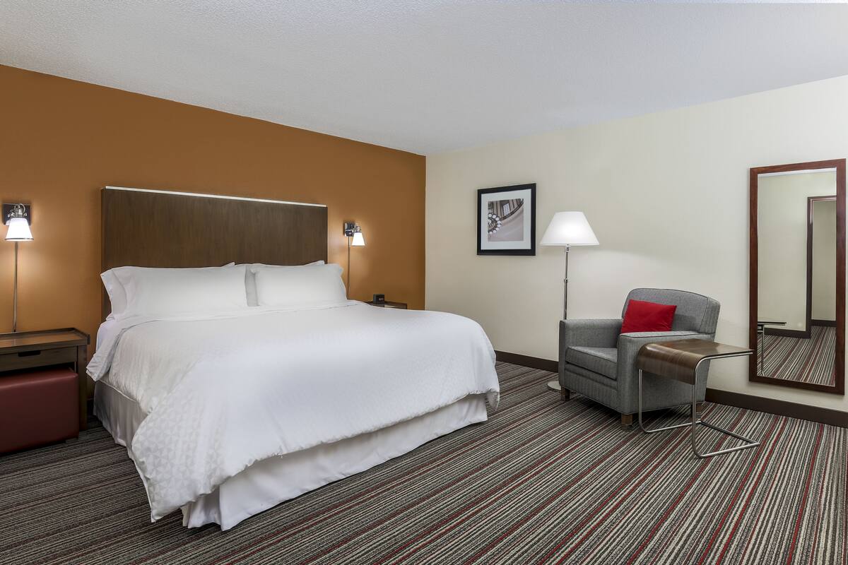A hotel room with a large bed, a chair, a desk, a lamp, and a framed picture on the wall.