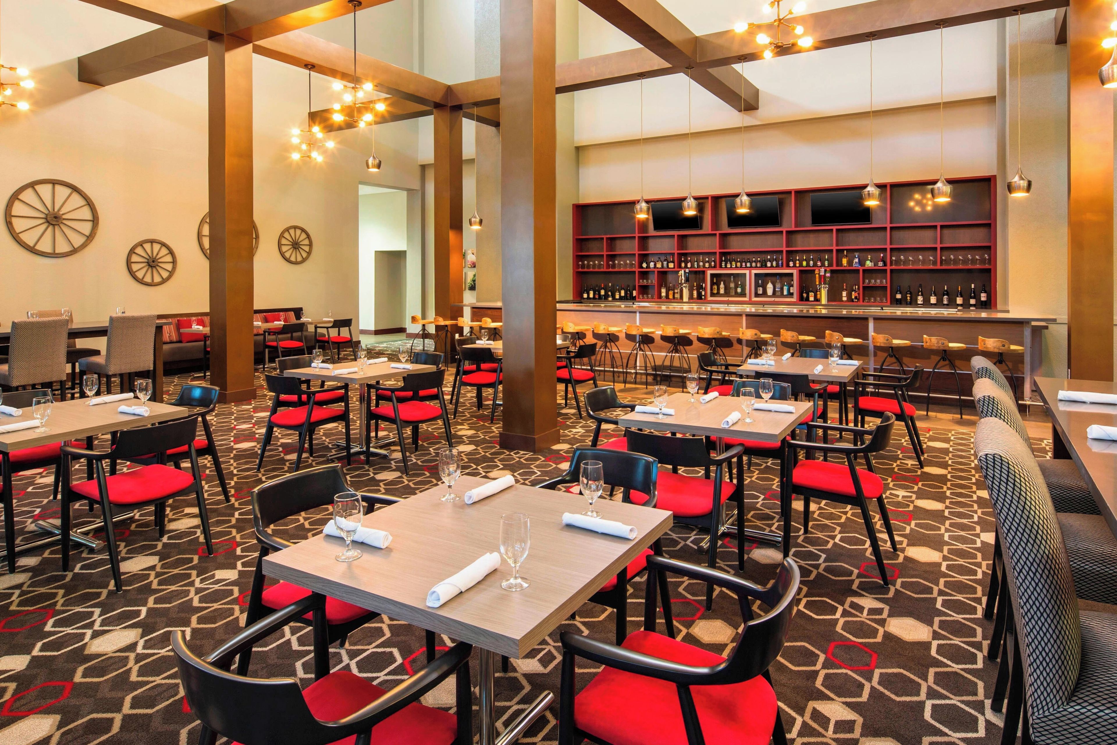 A hotel dining area with multiple tables and chairs, a bar with shelves of bottles, and a patterned carpet.