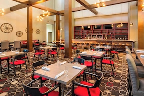 A hotel dining area with multiple tables and chairs, a bar with shelves of bottles, and a patterned carpet. - Four Points by Sheraton Bentonville (Bentonville)