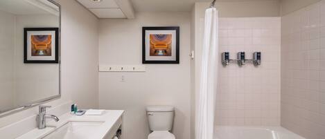 Une salle de bain avec un lavabo blanc, un miroir, des toilettes et une baignoire. Des tableaux encadrés ornent le mur, et il y a un rideau de douche.