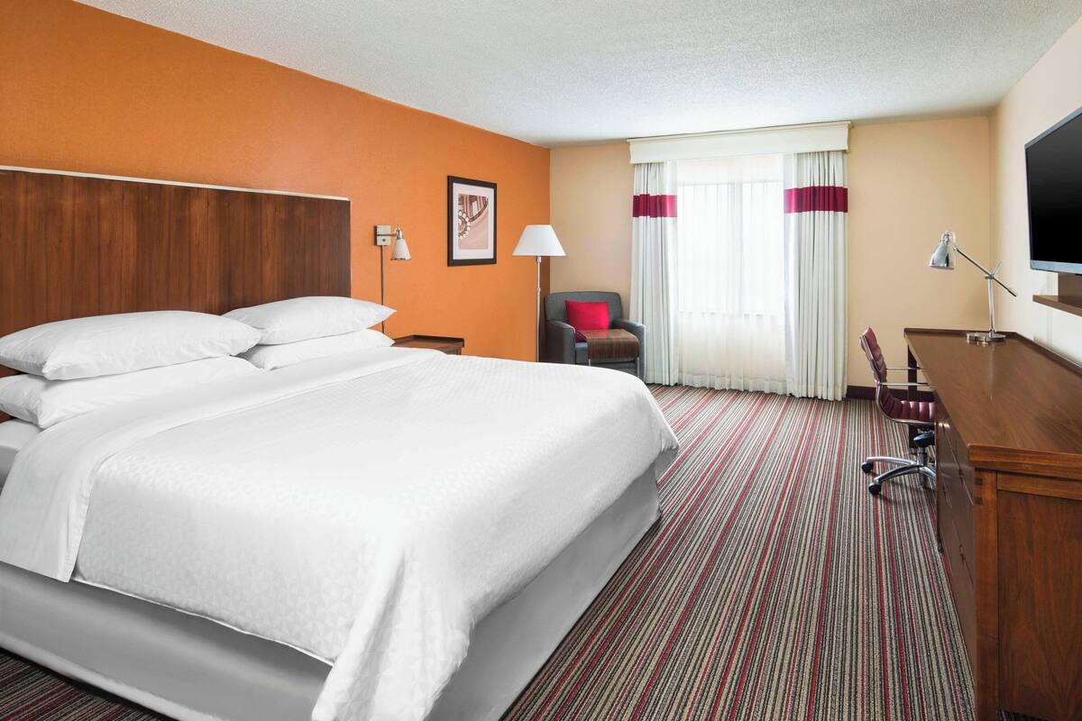 A hotel room with a large bed, a desk, a chair, a TV, and a window with curtains.