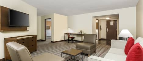 A hotel room with a flat-screen TV mounted on the wall, a desk with a chair, a sofa with red cushions, and a small table.