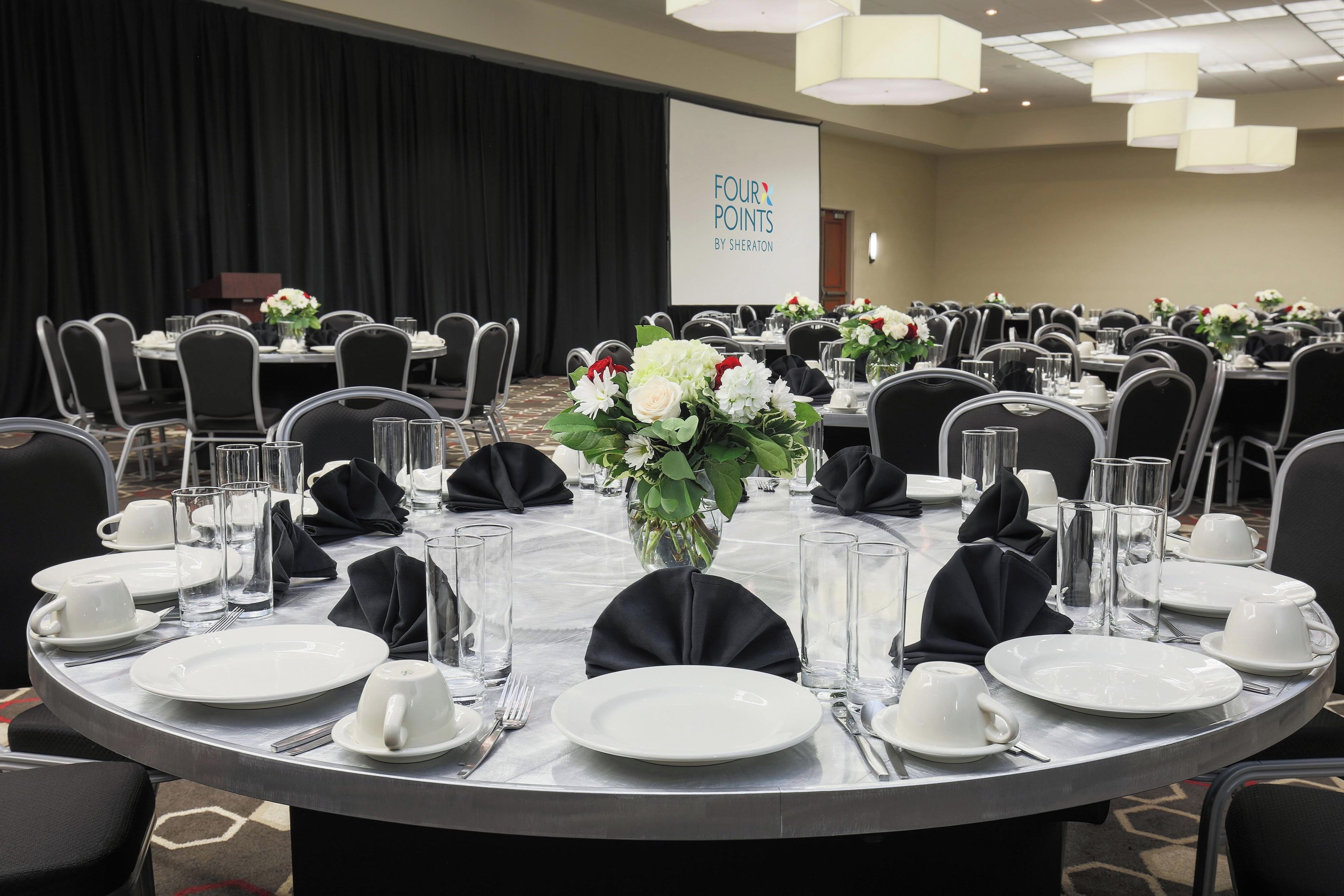 Un salón de conferencias con mesas redondas dispuestas para una reunión, con tarjetas de identificación, vasos de agua y centros de mesa florales.