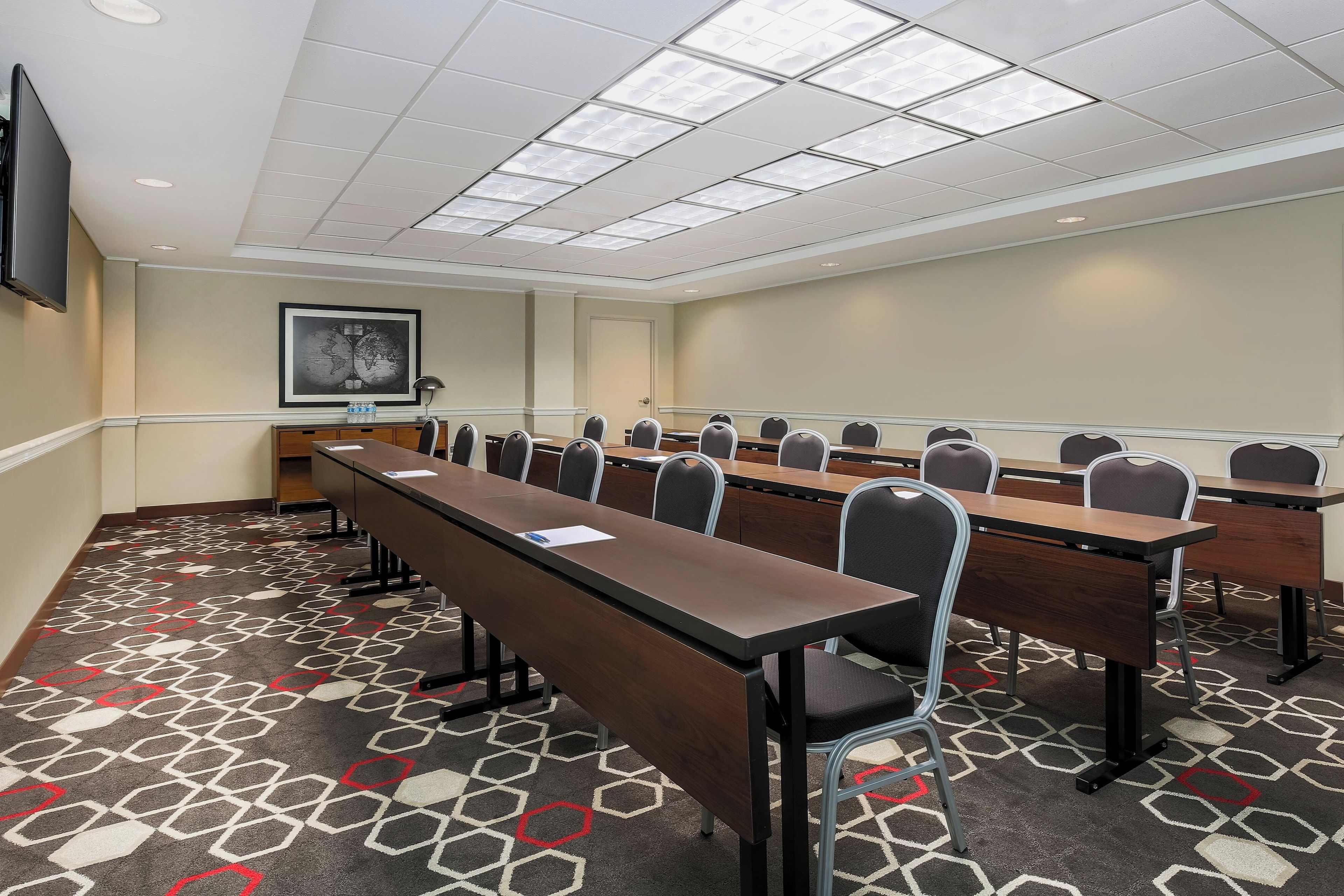A conference room with long tables and chairs, a framed picture on the wall, and a patterned carpet.