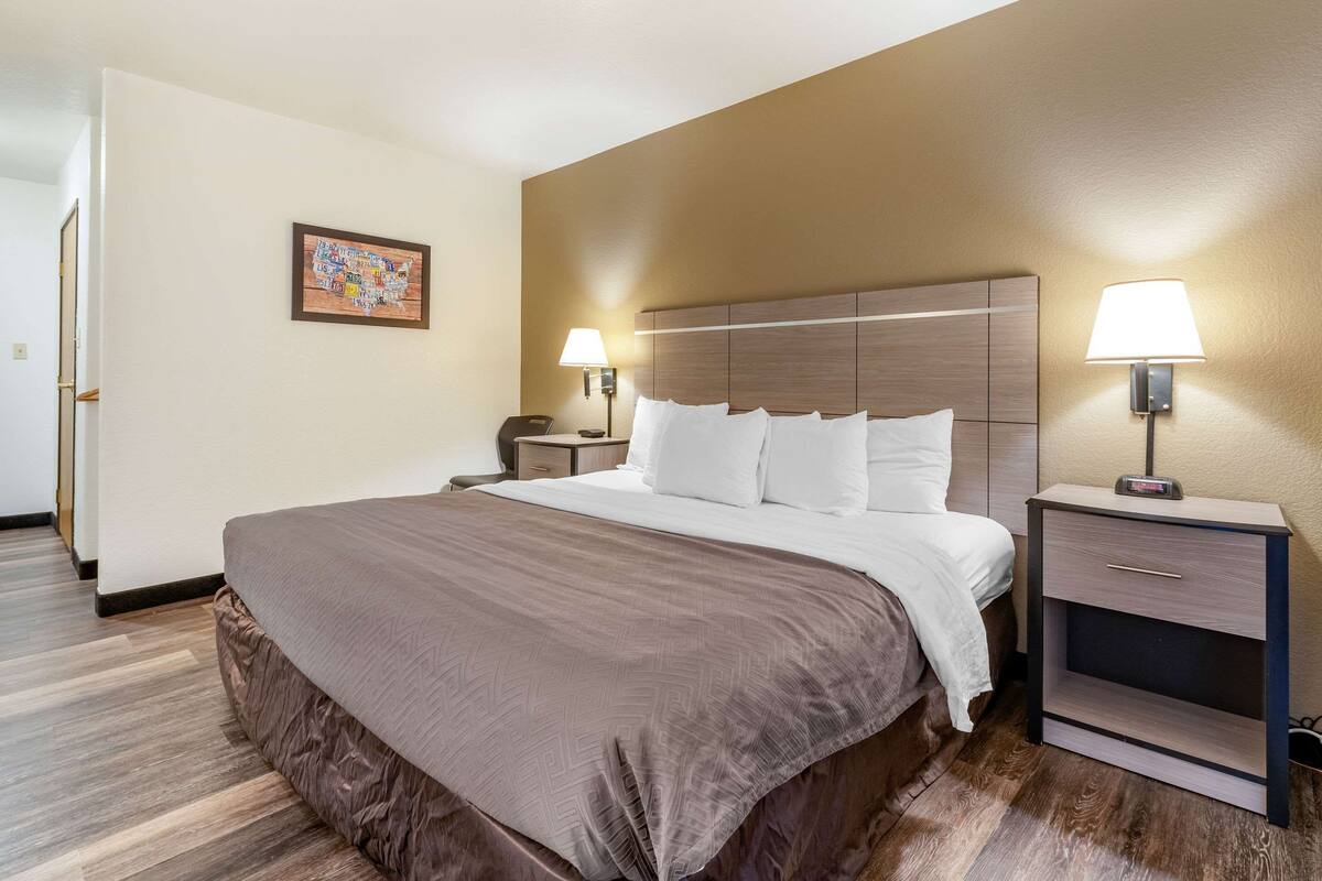 A hotel room with a large bed, two bedside tables with lamps, a nightstand, a wall-mounted clock, and a framed map on the wall.