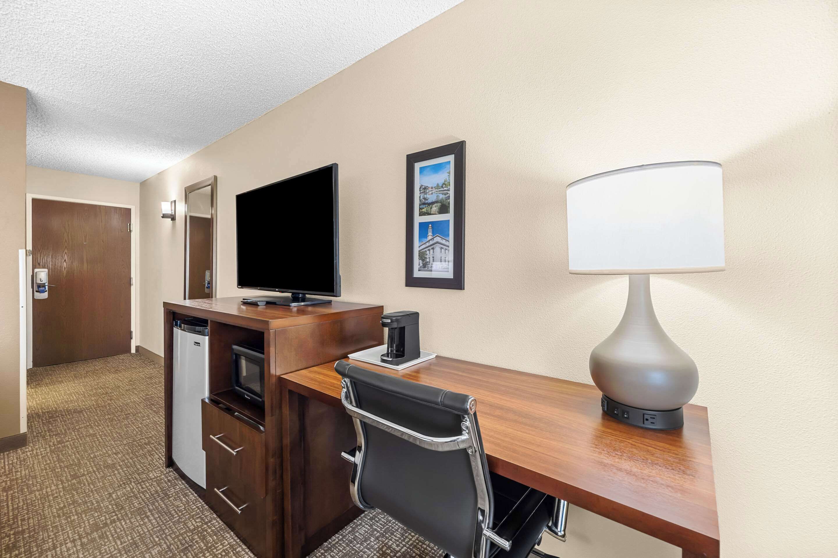 Habitación de hotel con un escritorio de madera, televisor, cafetera, lámpara, silla, refrigerador miniatura y un marco de fotos en la pared.