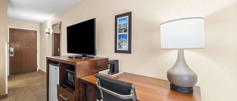 Habitación de hotel con un escritorio de madera, televisor, cafetera, lámpara, silla, refrigerador miniatura y un marco de fotos en la pared.