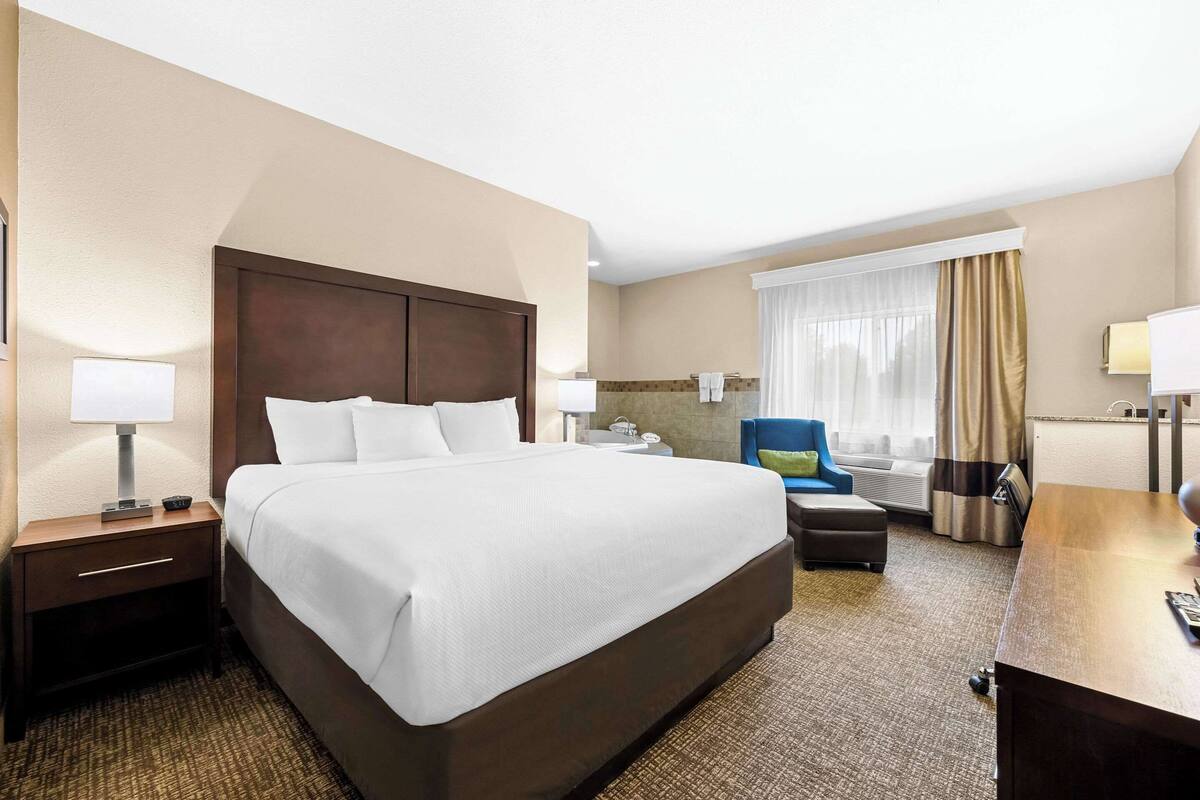 A hotel room with a large bed, a desk, a chair, a nightstand, and a window with curtains.