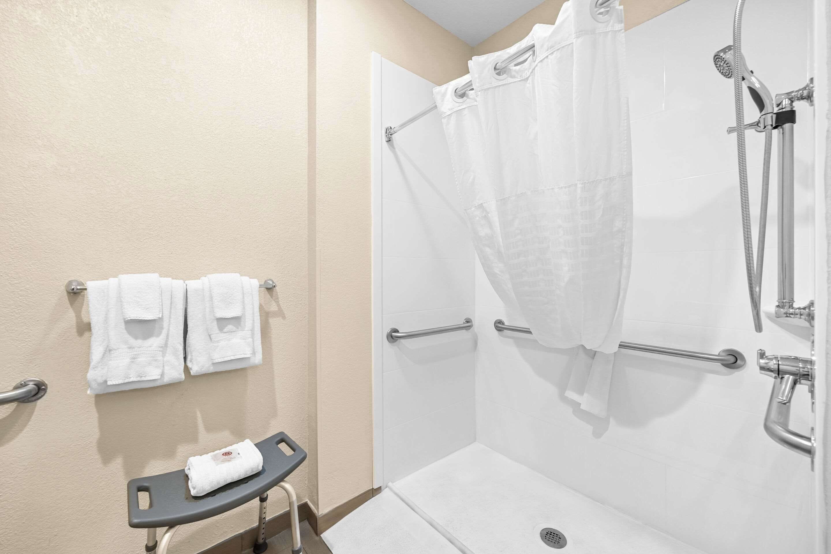 A shower area with a glass door, a showerhead, and a grab bar. There are two towels hanging on hooks and a towel on a small bench.