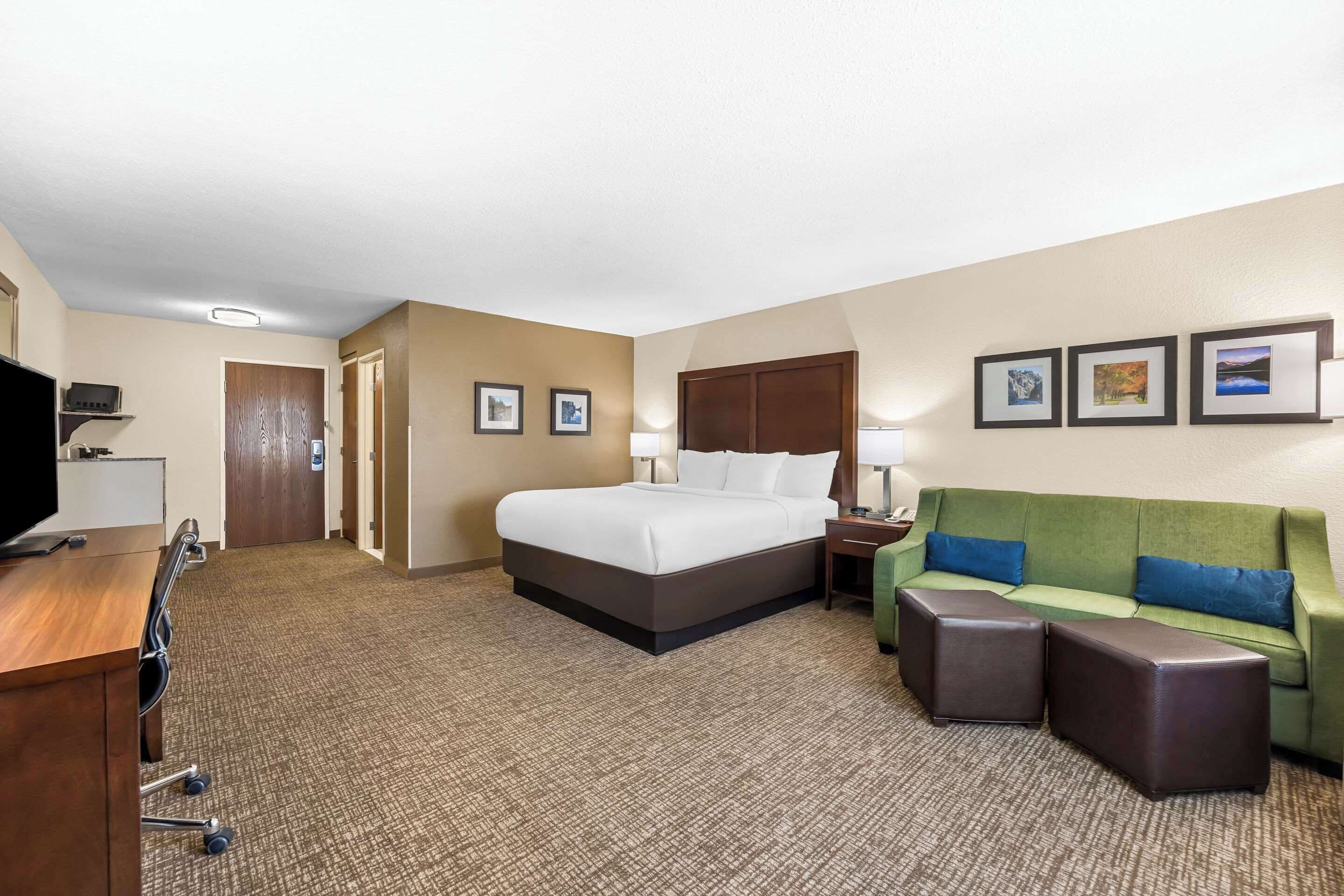 A hotel room with a bed, a green sofa, two ottomans, a desk with a television, and framed pictures on the wall.
