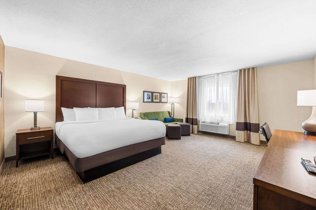 A hotel room with a large bed, a desk, a chair, a small table, and a window with curtains.