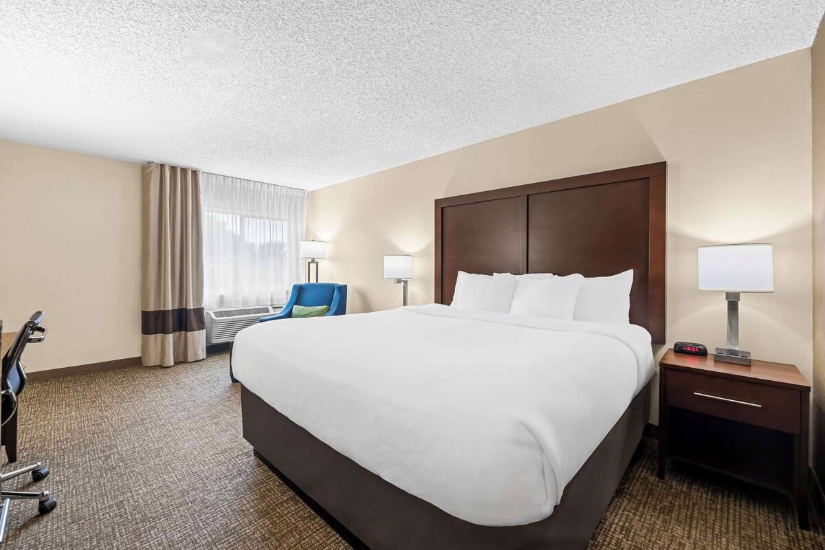 A hotel room with a large bed, two bedside tables with lamps, a desk with a chair, and a window with curtains.