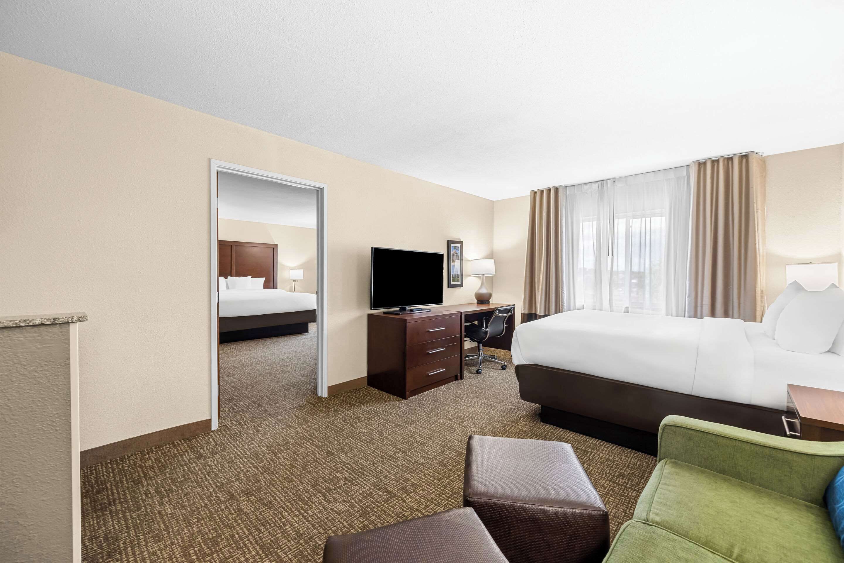 A hotel room with a bed, a desk with a computer, a television, and a seating area with a chair and ottoman.