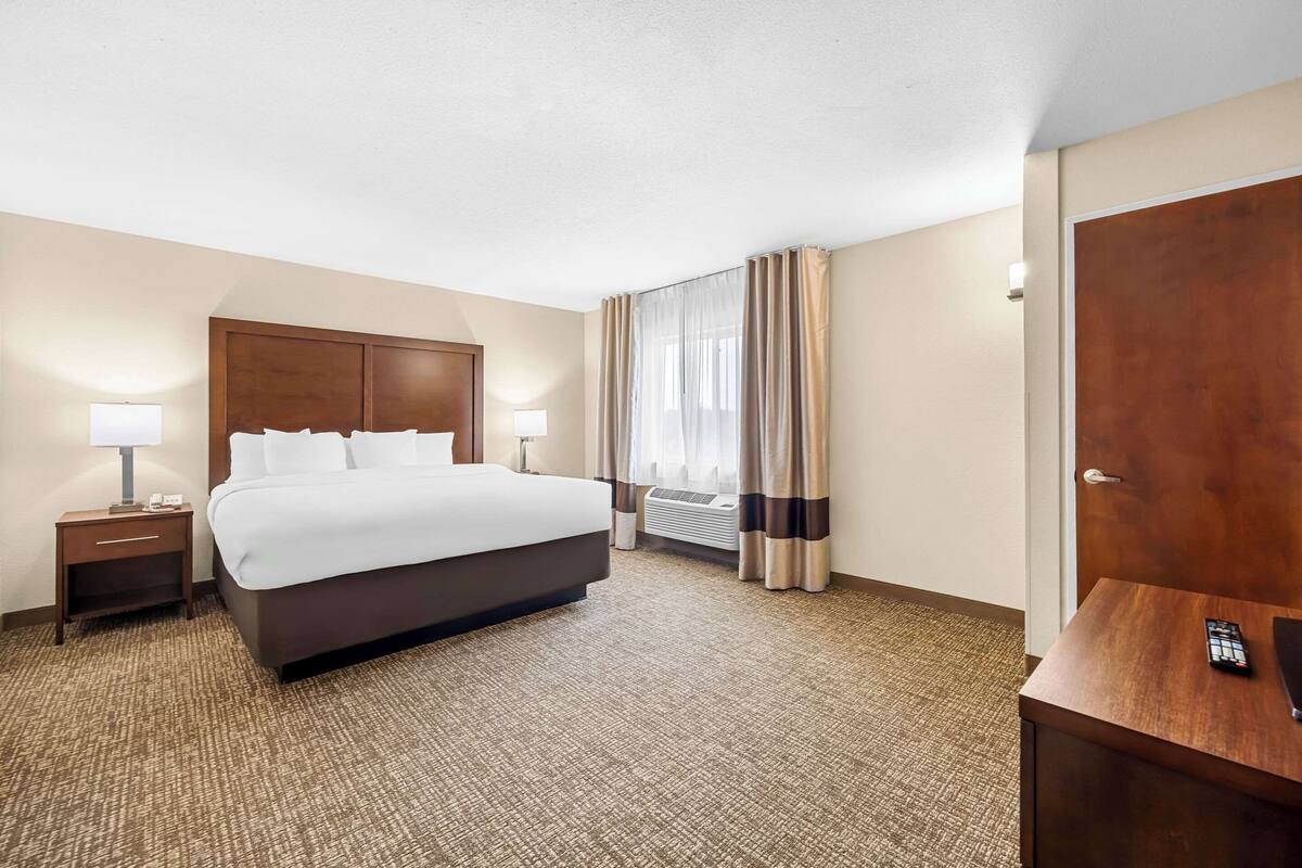 A hotel room with a large bed, a bedside table with a lamp, a desk with a remote control, and a door.