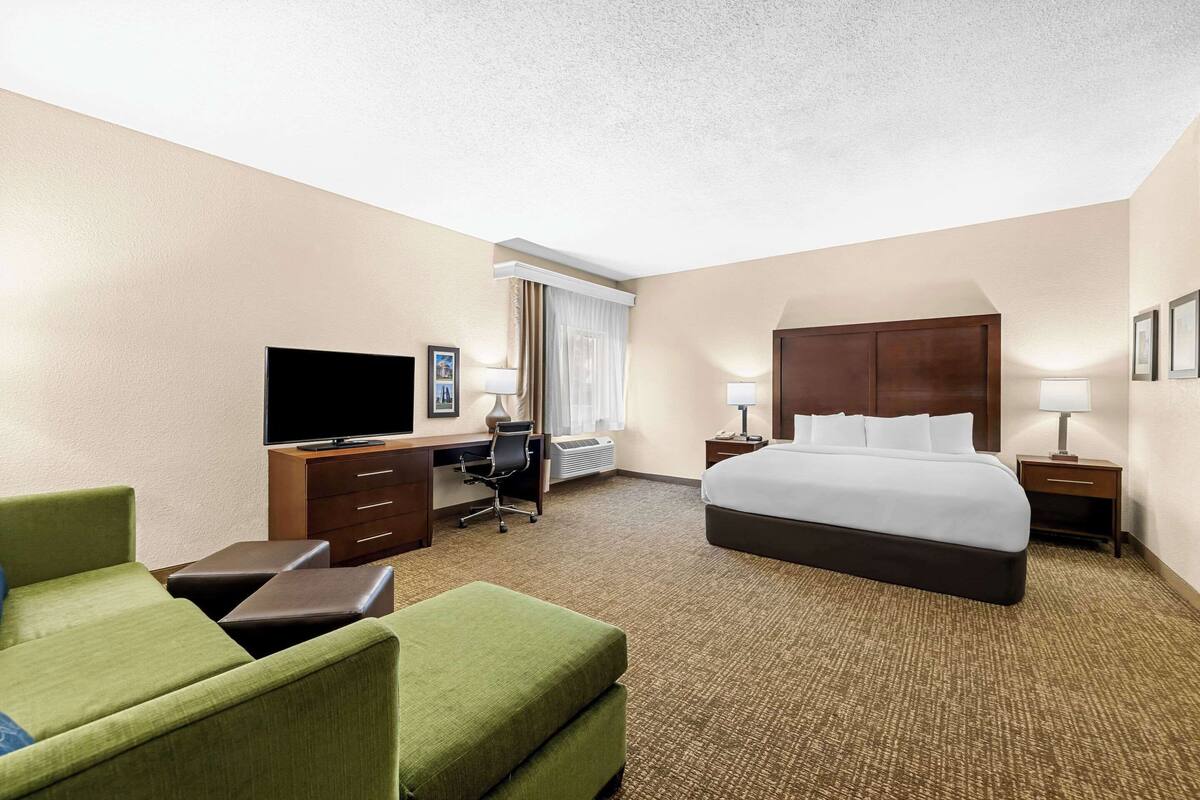 A hotel room with a bed, a desk with a chair, a television, and a seating area with ottomans.