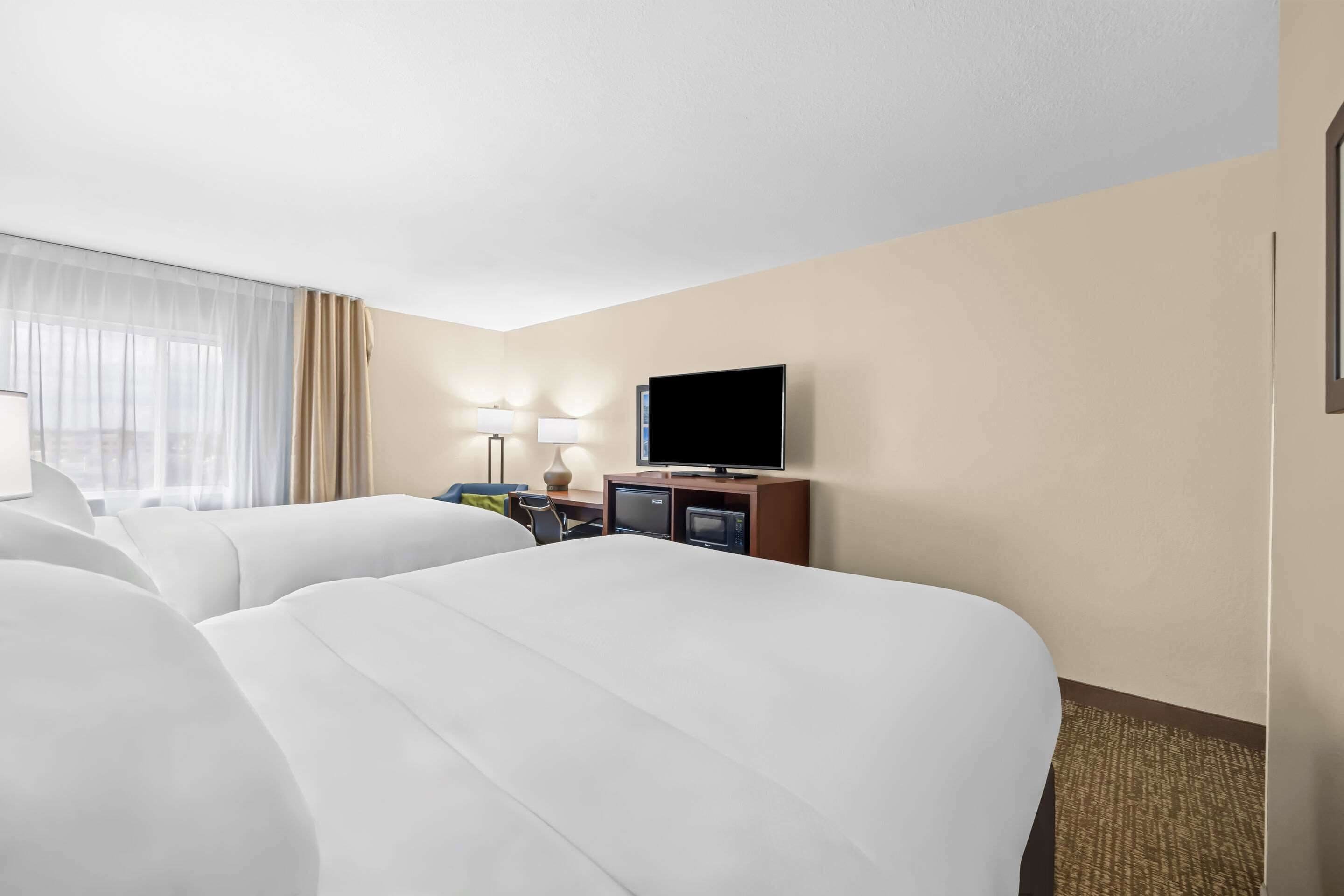A hotel room with two beds, a television, a desk with a lamp, and a view of the city through the window.