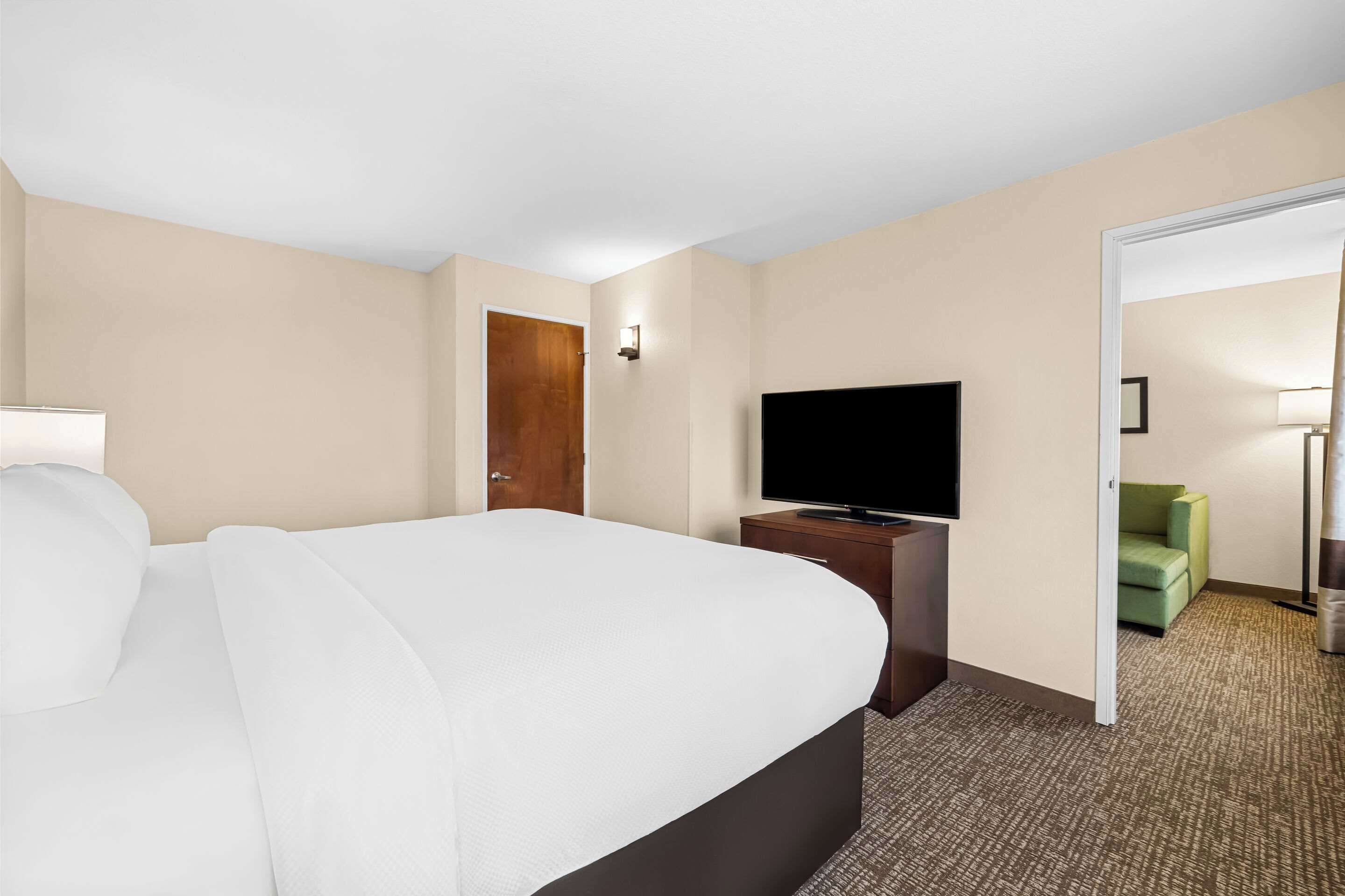 Ein Hotelzimmer mit einem großen Bett, einem Flachbildfernseher auf einem Holztischchen, einem grünen Sessel und einer Tür zu einem anderen Raum.