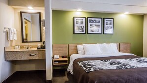 A hotel room with a bed, a nightstand, a vanity with a mirror, and a wall with framed pictures. - Rodeway Inn Austell Six Flags (Austell)