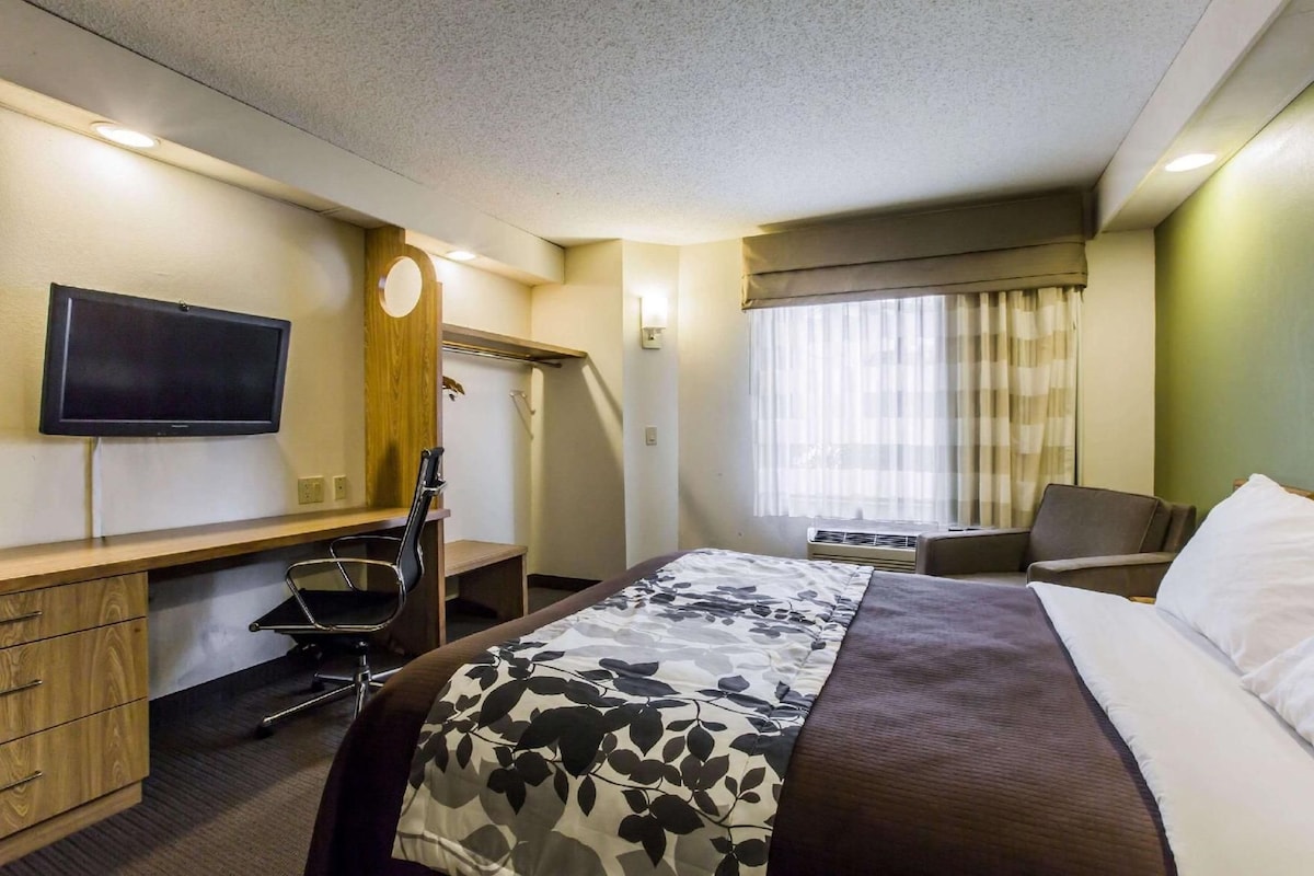Una habitación de hotel con cama, escritorio, silla, televisor y ventana con cortinas.