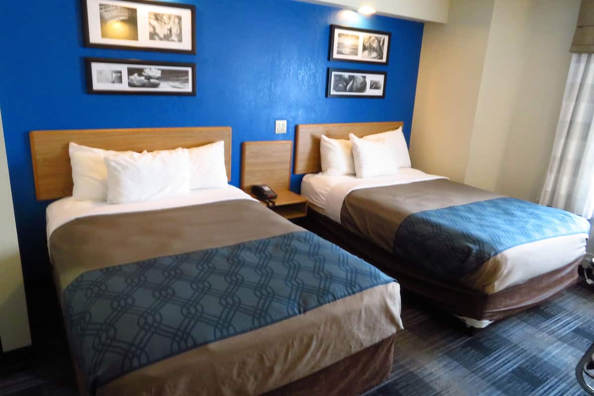 A hotel room with two beds, each with a wooden headboard, white bedding, and a patterned bedspread. There are framed pictures on the wall above the beds.