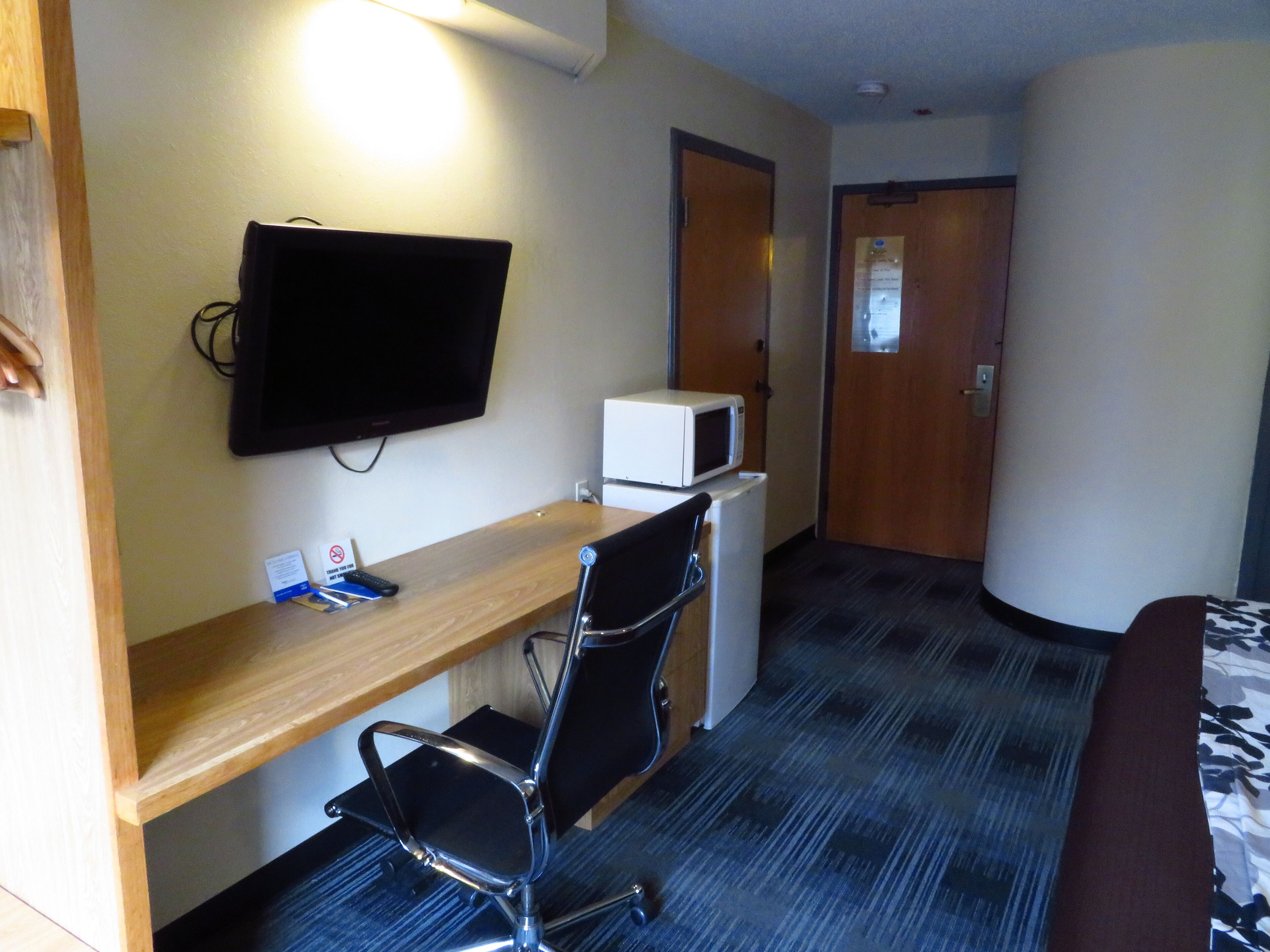 A hotel room with a wooden desk, a black office chair, a flat-screen TV mounted on the wall, a white microwave, and a refrigerator.