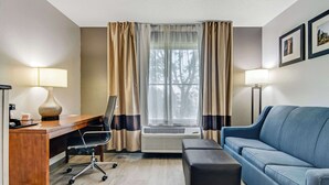 Habitación de hotel con escritorio de madera, una silla de oficina negra, sofá azul, reposapiés negro y una ventana con cortinas.