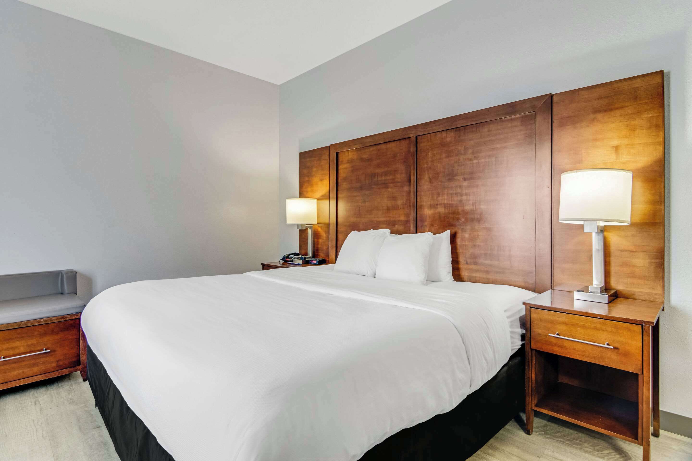 Habitación de hotel con una cama grande, dos mesitas de noche con lámparas, un banco y un cabecero de madera.