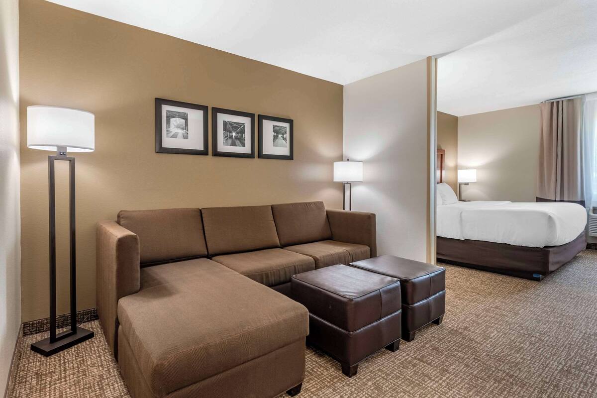 A hotel room with a brown sofa, two ottomans, a bed, and a floor lamp.