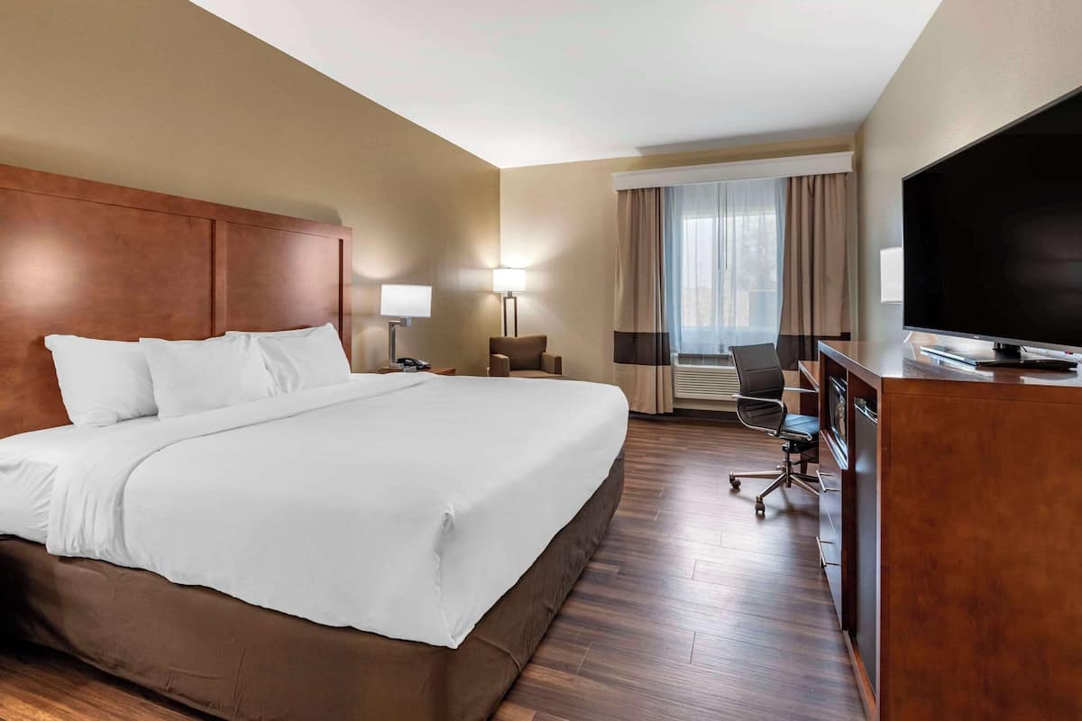 A hotel room with a large bed, a desk with a flat-screen TV, a chair, a lamp, and a window with curtains.