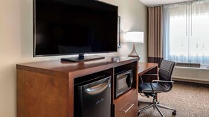 Une chambre d’hôtel équipée d’un téléviseur à écran plat, d’un bureau avec une lampe, d’un four à micro-ondes, d’un lave-vaisselle, d’un fauteuil de bureau noir et d’une grande fenêtre avec des rideaux.