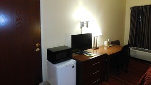 A compact hotel room with a wooden desk, a small white refrigerator, a television, a desk lamp, a chair, and a window with curtains. - Days Inn by Wyndham Walcott Davenport (Walcott)