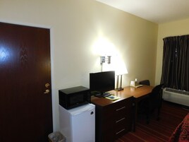 Una habitación de hotel compacta con un escritorio de madera, un refrigerador blanco pequeño, un televisor, una lámpara de escritorio, una silla y una ventana con cortinas.